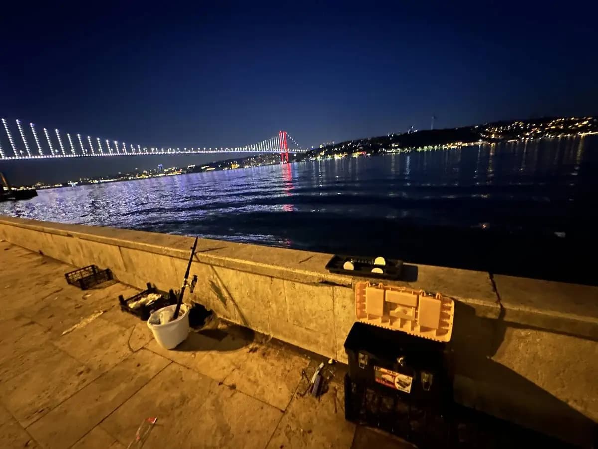 Rod, bucket and tackle box on the Kadıköy quay at night, the lit Bosphorus Bridge across the water